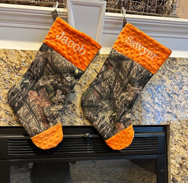 May include: Two personalized Christmas stockings with a camouflage pattern and orange trim. The stockings are hung on a fireplace mantel and are embroidered with the names "Jacob" and "Sawyer".