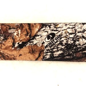 May include: Close-up of a brown, tan, and black camouflage fabric with the words "Mossy Oak Break-Up" printed in black.