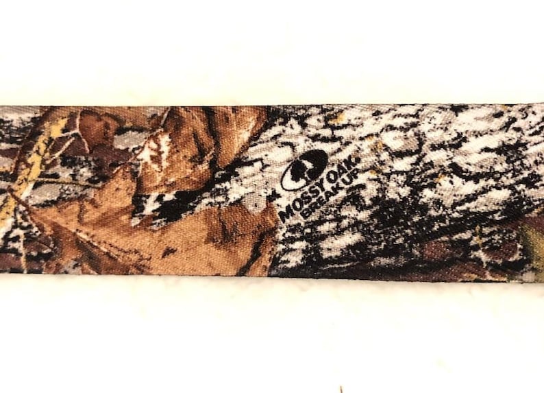 May include: Close-up of a brown, tan, and black camouflage fabric with the words "Mossy Oak Break-Up" printed in black.