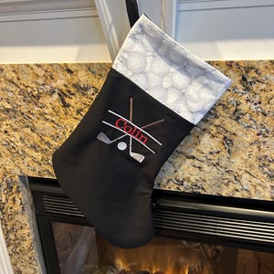May include: Black Christmas stocking with a golf theme. The stocking has a white cuff patterned with golf balls. The front features embroidered golf clubs, a golf ball, and the name "Colin" in red.