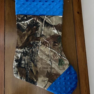 May include: A camouflage Christmas stocking with a blue fleece cuff and toe. The stocking is made of fabric and has a loop at the top for hanging.