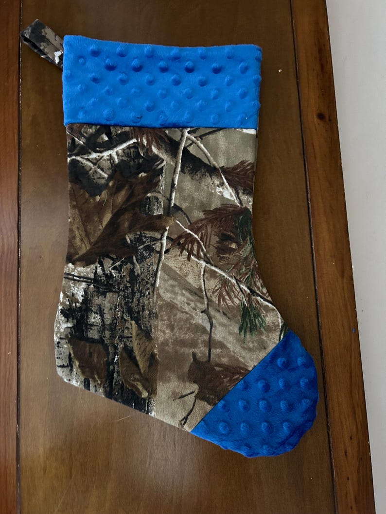 May include: A camouflage Christmas stocking with a blue fleece cuff and toe. The stocking is made of fabric and has a loop at the top for hanging.