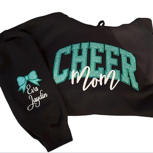 Puede incluir: Sudadera con capucha negra con el texto "Cheer Mom" en letras de purpurina turquesa. La manga izquierda tiene un lazo turquesa y el texto "Eva Jaydin" en blanco.
