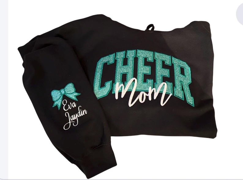 Puede incluir: Sudadera con capucha negra con el texto "Cheer Mom" en letras de purpurina turquesa. La manga izquierda tiene un lazo turquesa y el texto "Eva Jaydin" en blanco.