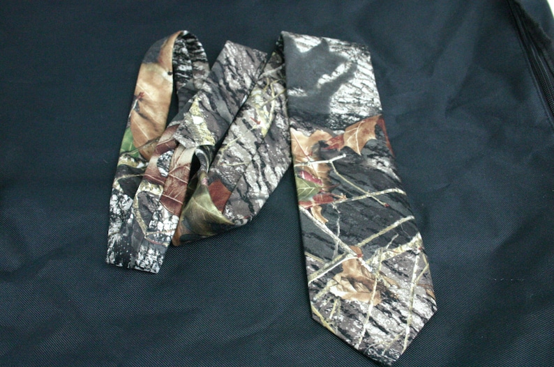 May include: A camouflage patterned tie with brown, green and white colours. The tie is made of a silky fabric and has a pointed tip.
