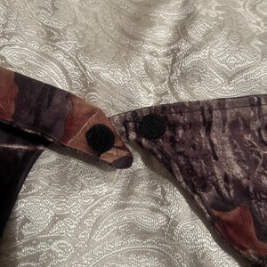 May include: Camouflage pattern fabric with two black hook-and-loop fasteners. The fabric features shades of brown, green, and black, creating a woodland camouflage design. The fasteners are circular and positioned to secure the fabric.
