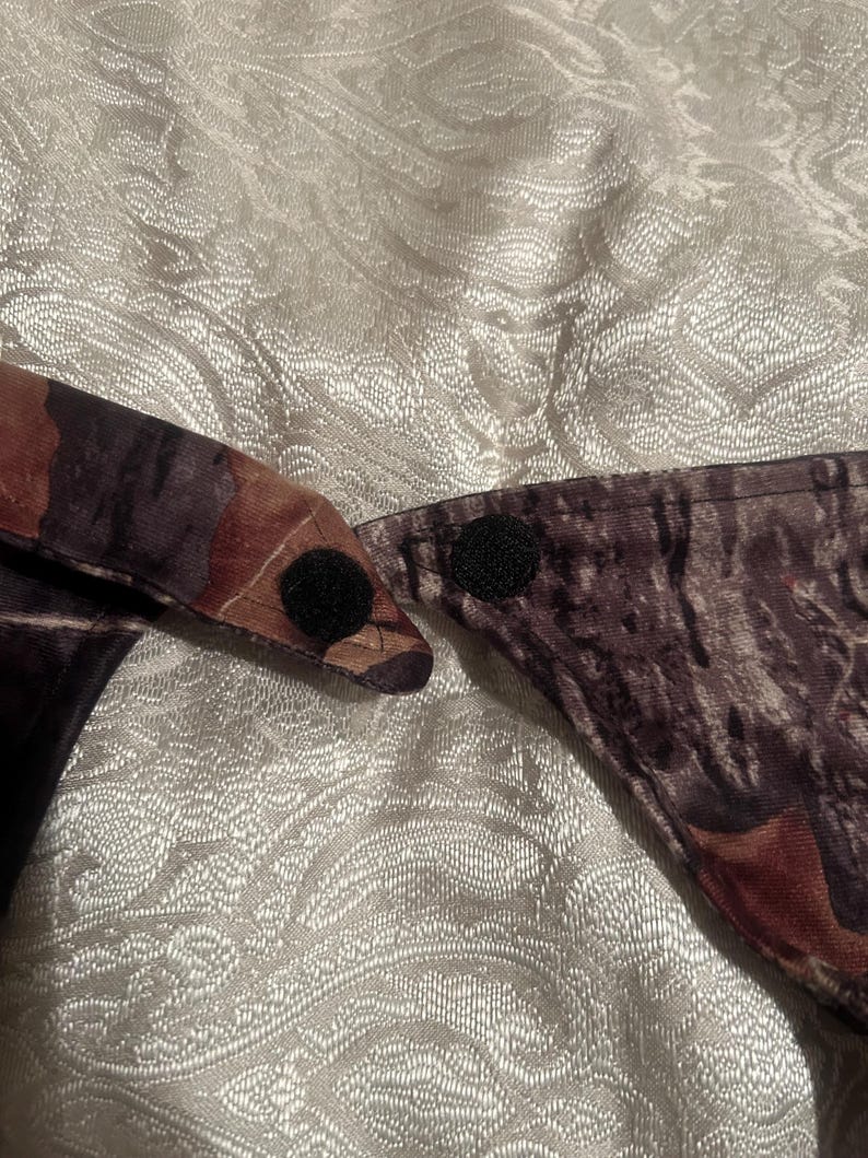May include: Camouflage pattern fabric with two black hook-and-loop fasteners. The fabric features shades of brown, green, and black, creating a woodland camouflage design. The fasteners are circular and positioned to secure the fabric.