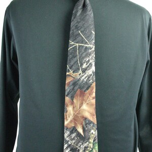 May include: A camouflage patterned tie with brown and green leaves. The tie is tied around a black shirt.