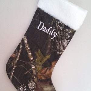 May include: Camouflage Christmas stocking with white plush trim and the word "Daddy" embroidered in white. The stocking features a woodland camouflage pattern with brown, green, and tan leaves and branches.