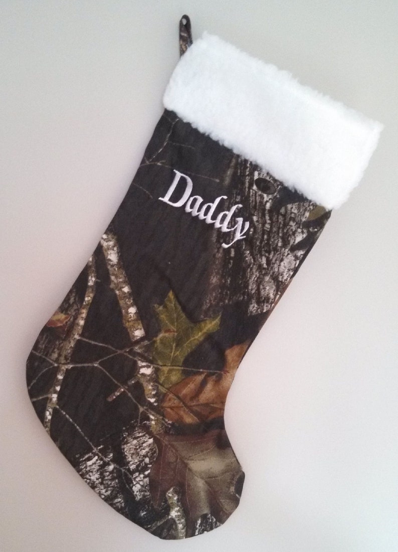 May include: Camouflage Christmas stocking with white plush trim and the word "Daddy" embroidered in white. The stocking features a woodland camouflage pattern with brown, green, and tan leaves and branches.