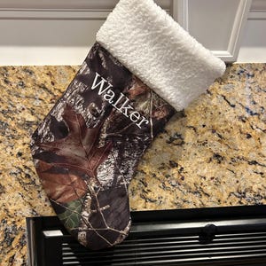 May include: Camouflage Christmas stocking with a white sherpa cuff. The name "Walker" is embroidered in white on the front. The stocking is hanging from a white hook.