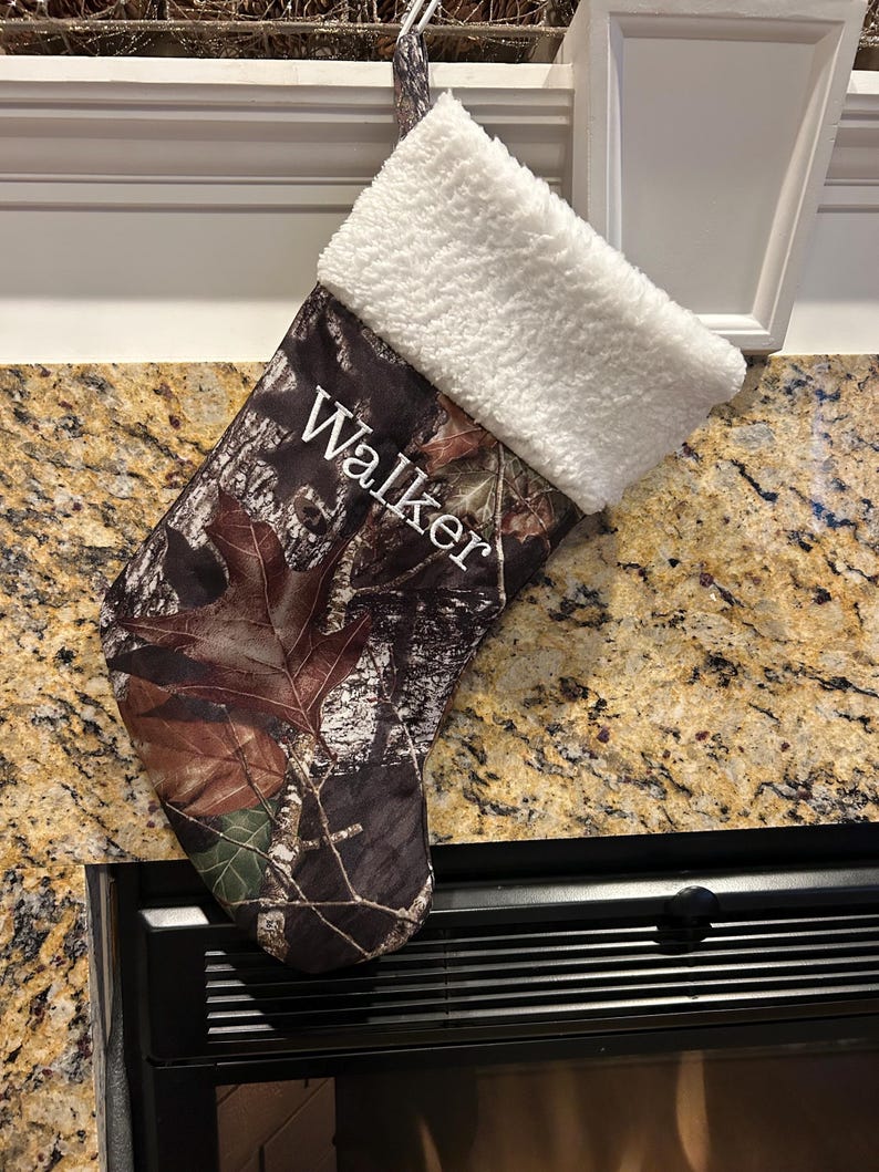 May include: Camouflage Christmas stocking with a white sherpa cuff. The name "Walker" is embroidered in white on the front. The stocking is hanging from a white hook.
