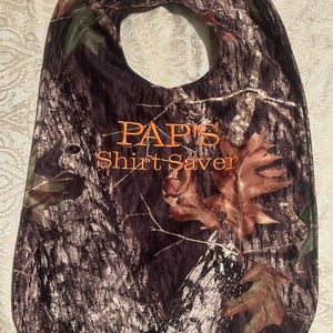 May include: Camouflage-patterned bib with the text "PAPI'S Shirt Saver" in orange. The bib features a large neck opening and is decorated with brown leaves.