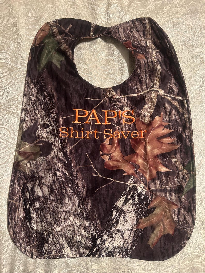 May include: Camouflage-patterned bib with the text "PAPI'S Shirt Saver" in orange. The bib features a large neck opening and is decorated with brown leaves.