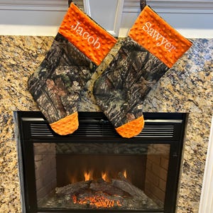 May include: Two personalized Christmas stockings with a camouflage pattern and orange felt trim. The stockings are hung on a white mantel above a fireplace with a lit fire.