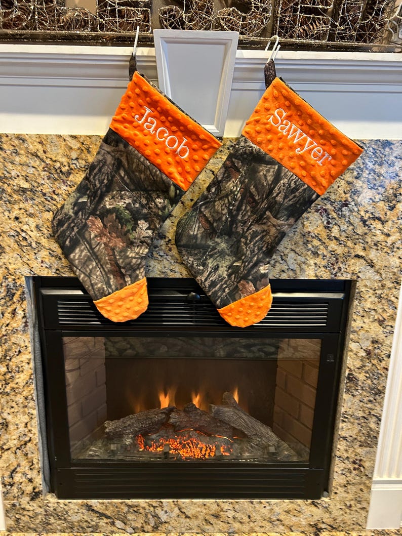 May include: Two personalized Christmas stockings with a camouflage pattern and orange felt trim. The stockings are hung on a white mantel above a fireplace with a lit fire.