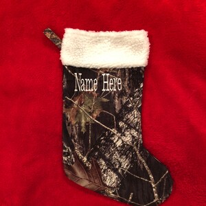 May include: A Christmas stocking with a camouflage pattern, featuring a white fluffy cuff and a loop for hanging. The text "Name Here" is embroidered in white. The stocking is set against a red background.