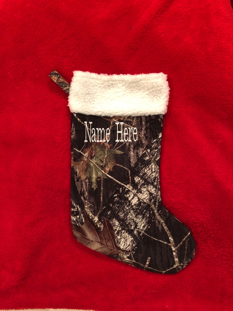 May include: A Christmas stocking with a camouflage pattern, featuring a white fluffy cuff and a loop for hanging. The text "Name Here" is embroidered in white. The stocking is set against a red background.