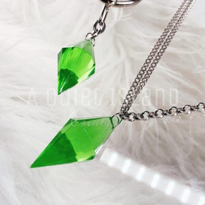 May include: A silver-toned metal chain necklace featuring two faceted, diamond-shaped green crystal pendants. The pendants are suspended from a metallic link chain. The text A quiet island creative is visible in the background behind the jewelry.