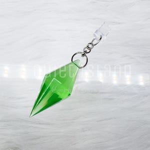 May include: A translucent, bright green, diamond-shaped pendant with faceted edges, attached to a silver-toned metal jump ring and a short chain link. The pendant features a sharp, pointed bottom and a wider, angular middle section.
