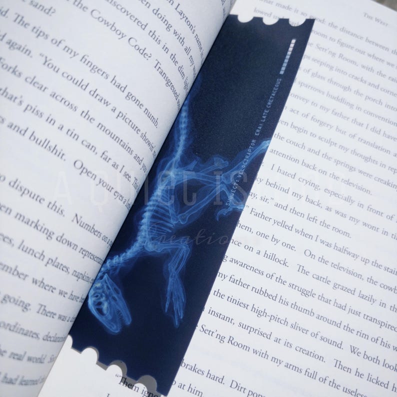 May include: A bookmark featuring a blue X-ray image of a Velociraptor skeleton. The bookmark has a dark blue background with white text that reads "LUQUECT: VELOCIRAPTOR CRAI LATE CRETACEOUS". The bookmark is placed between the pages of a book.