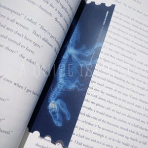 May include: A blue bookmark featuring a detailed, glowing x-ray image of a dinosaur skeleton. The bookmark is rectangular with a scalloped edge and is placed between the pages of a book.