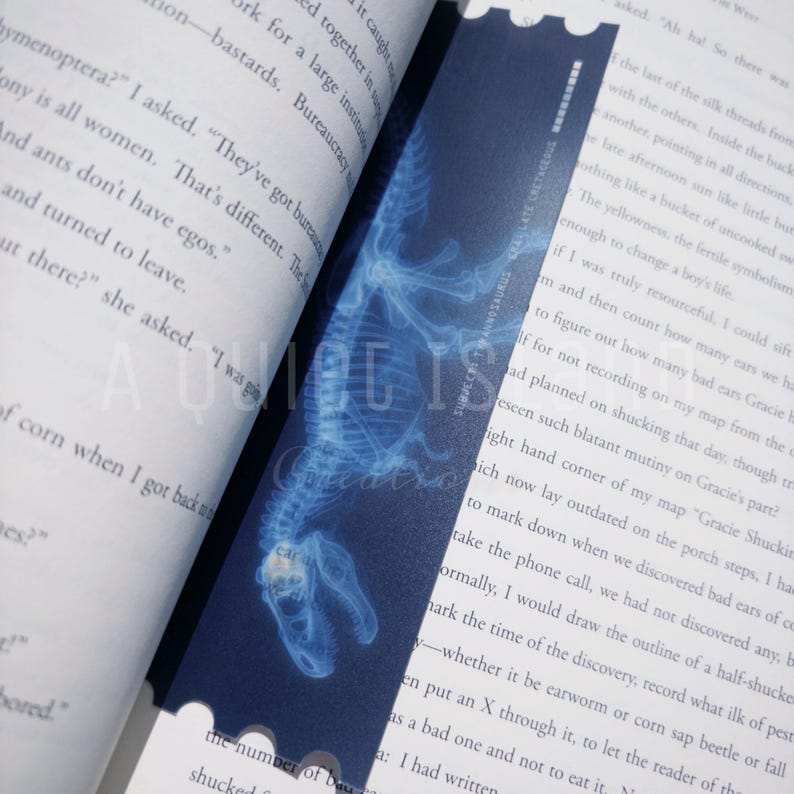 May include: A blue bookmark featuring a detailed, glowing x-ray image of a dinosaur skeleton. The bookmark is rectangular with a scalloped edge and is placed between the pages of a book.
