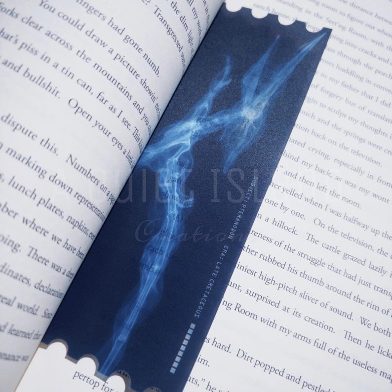 May include: A dark blue bookmark featuring a detailed, glowing blue illustration of a pteranodon fossil. The bookmark has a scalloped edge and is placed between the pages of a book. Text on the bookmark reads "SUBJECT: PTERANODON ERA: LATE CRETACEOUS."
