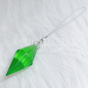 May include: A bright green, translucent, diamond-shaped pendant charm with a faceted surface. The charm is attached to a silver-toned metal jump ring and a small, textured silver bead, connected to a thin, light gray cord loop for hanging.