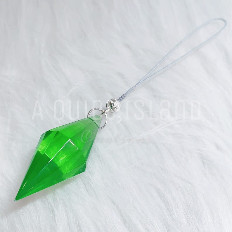 May include: A bright green, translucent, diamond-shaped pendant charm with a faceted surface. The charm is attached to a silver-toned metal jump ring and a small, textured silver bead, connected to a thin, light gray cord loop for hanging.