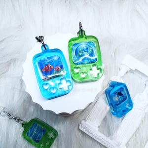 May include: Four resin keychains shaped like handheld gaming consoles. Two are blue with clownfish and orca graphics, and two are green with abstract blue circular designs. Each features a metal jump ring for attachment and a glossy finish.