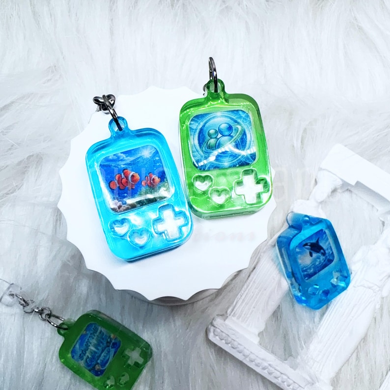 May include: Four resin keychains shaped like handheld gaming consoles. Two are blue with clownfish and orca graphics, and two are green with abstract blue circular designs. Each features a metal jump ring for attachment and a glossy finish.