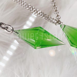 May include: Two translucent lime green diamond-shaped pendants attached to silver-toned metal chains. The pendants have a faceted, geometric design. The text A QUIET ISLAND CREATIONS is overlaid in white across the center of the image.