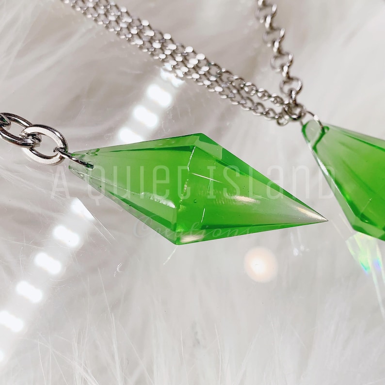 May include: Two translucent lime green diamond-shaped pendants attached to silver-toned metal chains. The pendants have a faceted, geometric design. The text A QUIET ISLAND CREATIONS is overlaid in white across the center of the image.
