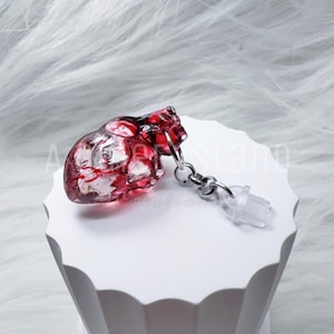 May include: A clear resin heart-shaped keyring with red detailing, connected to a silver-coloured chain and a clear plastic clasp. The heart features internal structures. The keyring is displayed on a white, textured surface.