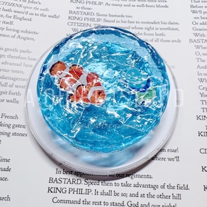 May include: A circular, translucent blue resin coaster featuring a textured, water-like surface. Embedded within the resin is a small, orange and white clownfish figure and a tiny green sea turtle, creating an underwater scene effect.