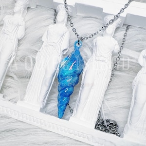 May include: A blue seashell pendant necklace with a silver chain. The pendant is a vibrant blue with glitter accents. The necklace is displayed against a backdrop of white statues.