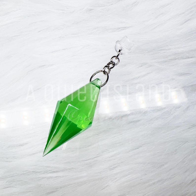 May include: A translucent, bright green, diamond-shaped pendant with faceted edges. The pendant is attached to a small silver-toned metal jump ring and a short chain link, which connects to a clear, cylindrical plastic cap at the top end.