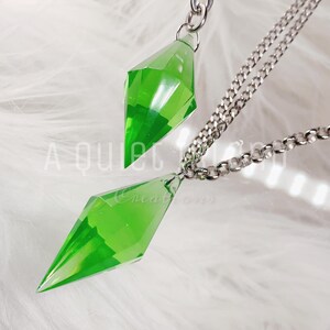 May include: Two translucent, bright green, diamond-shaped pendants hanging from silver-toned metal chains. The pendants have a faceted, geometric design that tapers to a point at both ends. The text A QUIET ISLAND Creations is visible.