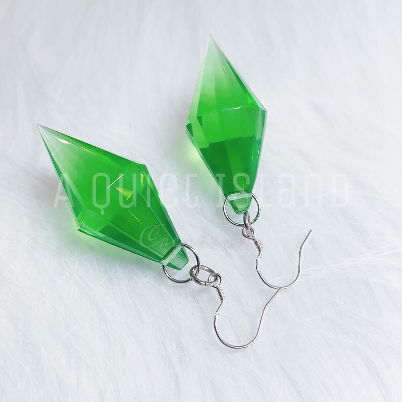 May include: A pair of dangle earrings featuring translucent, bright green, diamond-shaped geometric pendants. Each pendant is attached to a silver-toned metal fishhook ear wire by a small circular jump ring. The design is faceted and pointed.