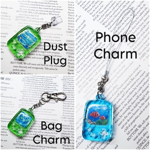 May include: A collection of rectangular resin charms featuring underwater scenes with clownfish. The set includes a green dust plug, a green bag charm with a metal lobster clasp, and a blue phone charm with a thin cord loop attachment.