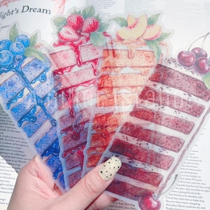 May include: A hand holds four transparent stickers shaped like cake slices, each topped with different fruits. The cakes are decorated with blueberries, strawberries, peaches, and cherries. The stickers are layered over a printed page.