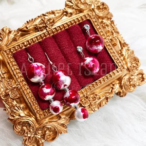 May include: Dangle earrings with red and white marbled spheres. The earrings are displayed on a red velvet surface inside a gold ornate frame. The earrings have silver-colored hooks and small clear gems.