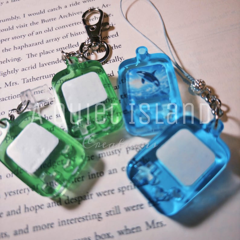 May include: Four translucent plastic handheld game keychains in green and blue. Each features a rectangular screen area, a D-pad, and buttons. One blue keychain displays an orca graphic. The keychains include metal lobster clasps or a cord loop.