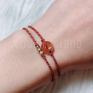 May include: Two red and brown string bracelets with a round, light brown gemstone charm. The bracelets are tied together with a gold bead.