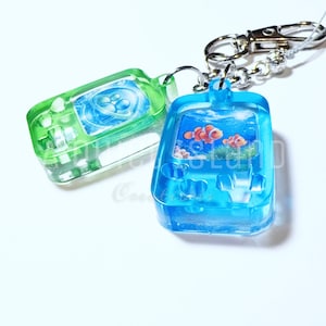 May include: Two translucent resin keychains shaped like handheld gaming consoles. One is lime green with a blue circular graphic, and the other is bright blue featuring an underwater scene with two orange clownfish. Both have metal hardware.