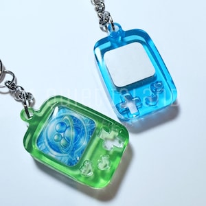 May include: Two translucent resin keychains shaped like handheld gaming consoles. One is lime green with a blue circular graphic on the screen, and the other is bright blue with a blank white screen. Both feature silver metal key rings.