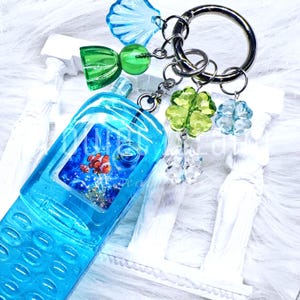 May include: A blue resin keyring shaped like a vintage mobile phone with a screen displaying a clownfish. The keyring features a silver metal ring adorned with charms including a blue seashell, a green bell, and three faceted clover-shaped beads.