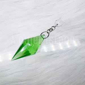 May include: A translucent green diamond-shaped pendant with faceted edges, attached to a silver-toned metal jump ring and a clear plastic connector. The object features a sharp, pointed base and a geometric design, reflecting light across its surface.