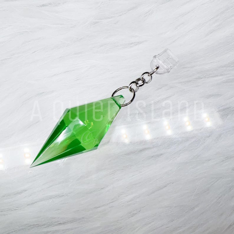 May include: A translucent green diamond-shaped pendant with faceted edges, attached to a silver-toned metal jump ring and a clear plastic connector. The object features a sharp, pointed base and a geometric design, reflecting light across its surface.
